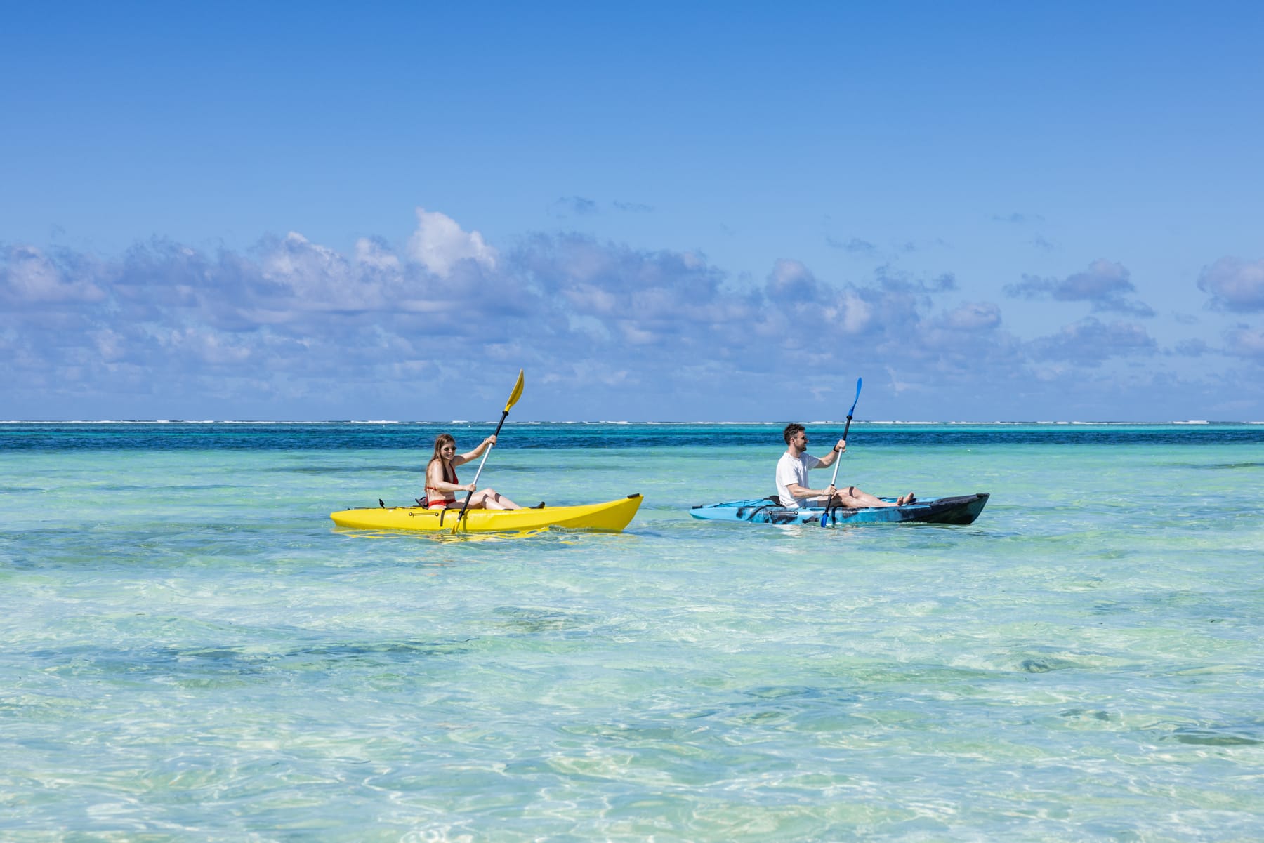 Two people paddling a yellow and a blue kayak across the clear turquoise lagoon in front of Matlai